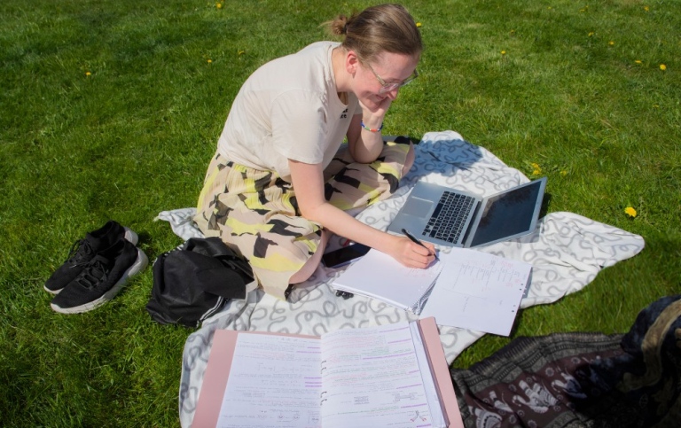 En kvinna sitter i gräset på en filt med en dator, papper och penna.